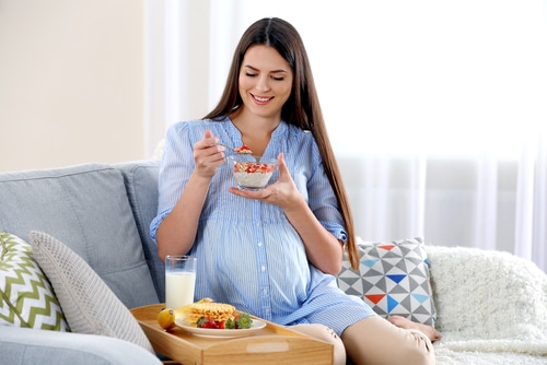 Young pregnant woman with milk and breakfast on sofa